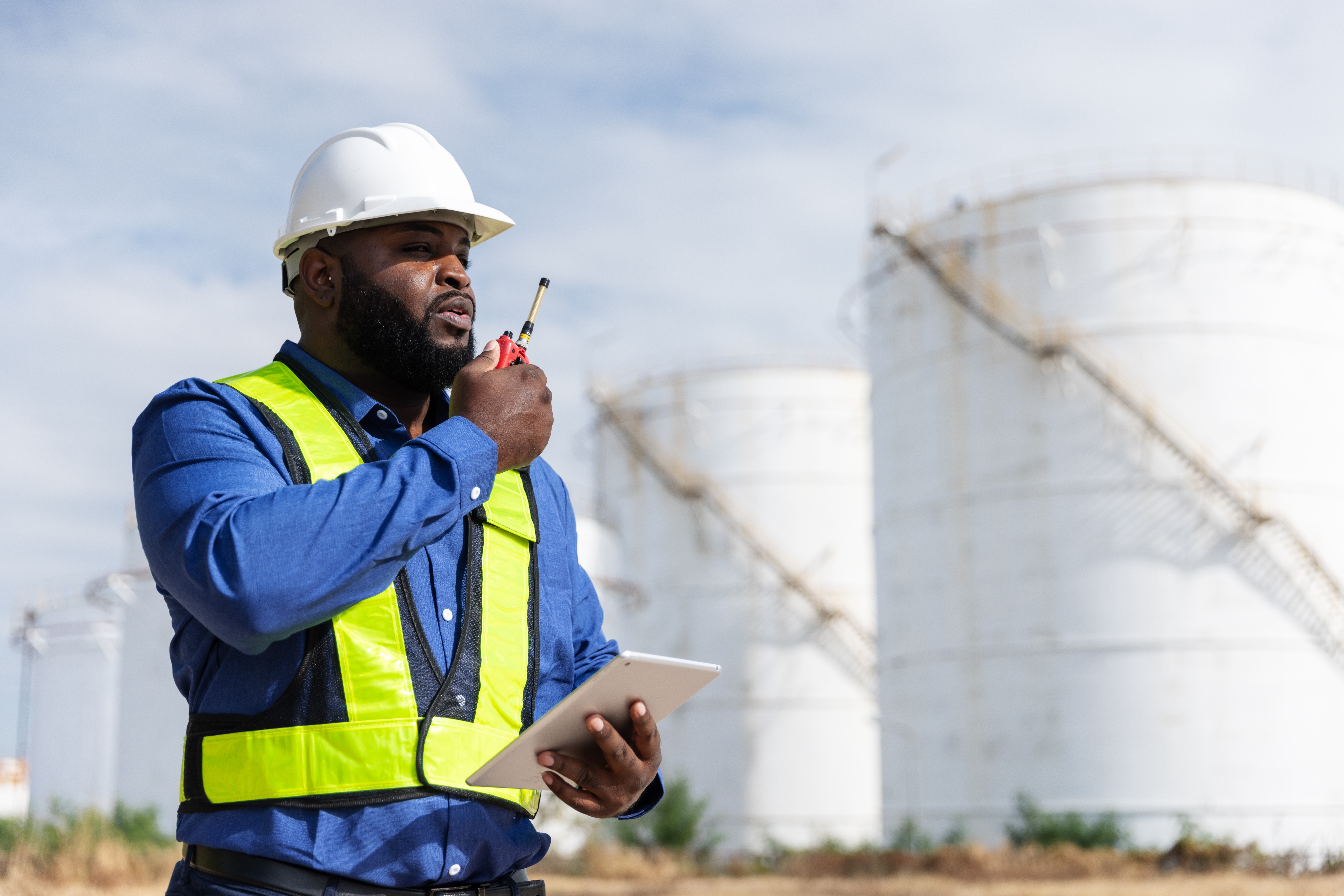 A safety engineer wearing a white hard hat, blue shirt, and yellow reflective vest uses a two way radio while holding a tablet. The background features large industrial storage tanks under a blue sky